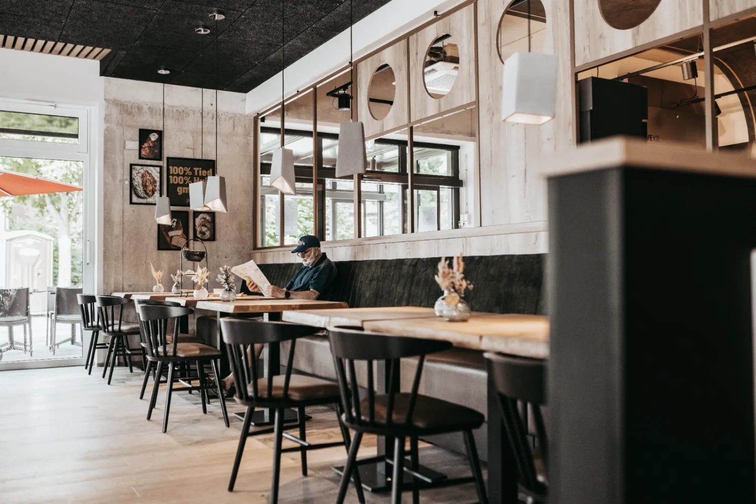 An einer verspiegelten Wand ist eine lange Sitzbak mit mehreren Tischen und Stühlen. Ein Mann sitzt dort und liesst Zeitung.